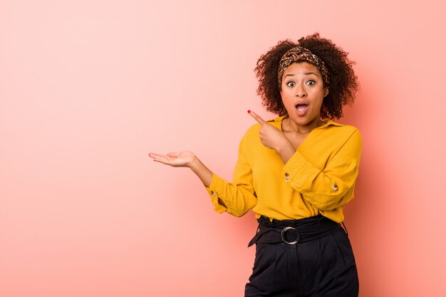 La giovane donna afroamericana contro una parete rosa ha eccitato tenendo uno spazio della copia sul palmo.