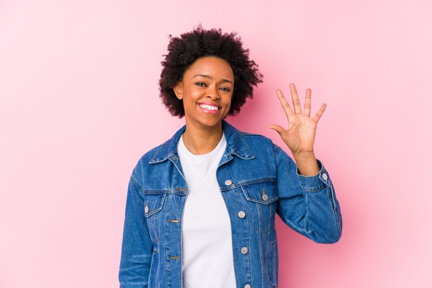 La giovane donna afroamericana contro una parete rosa ha isolato il numero di mostra allegro sorridente sorridente cinque con le dita.