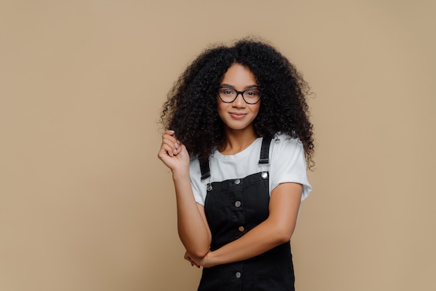 La giovane donna afroamericana dall'aspetto piacevole tocca i capelli ricci, ha un'acconciatura folta, indossa occhiali da vista