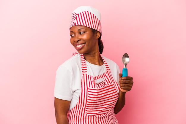 La giovane donna afroamericana del gelatiere che tiene la paletta isolata su sfondo rosa guarda da parte sorridente, allegra e piacevole.