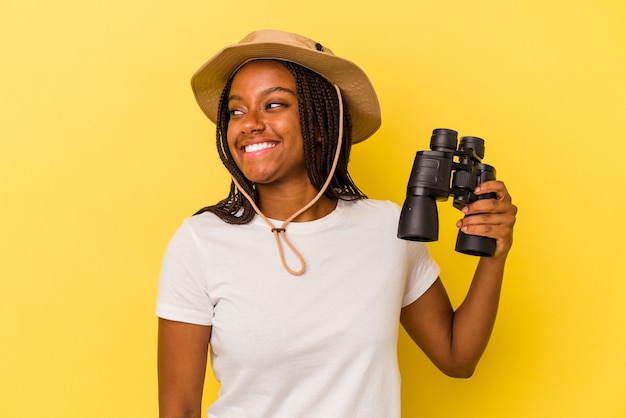 La giovane donna afroamericana dell'esploratore che tiene un binocolo isolato su fondo giallo guarda da parte sorridente, allegra e piacevole.