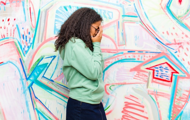 La giovane donna afroamericana graziosa che copre gli occhi di mani con uno sguardo triste e frustrato di disperazione, gridando contro la parete dei graffiti