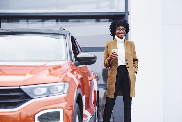 La giovane donna afroamericana in vetri e con la tazza della bevanda sta all'aperto vicino all'automobile moderna.
