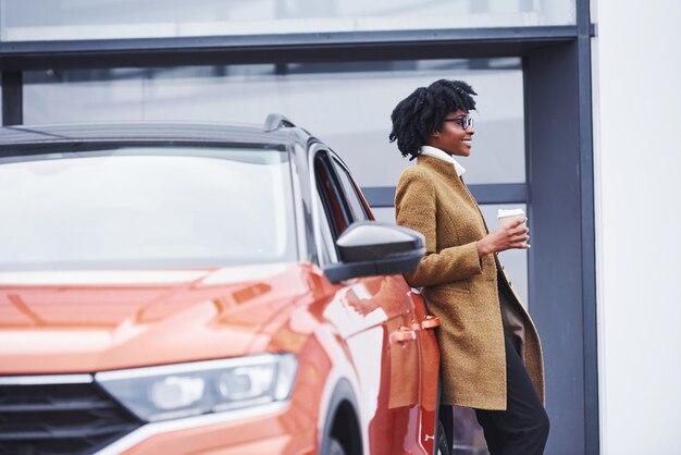 La giovane donna afroamericana in vetri e con la tazza della bevanda sta all'aperto vicino all'automobile moderna.