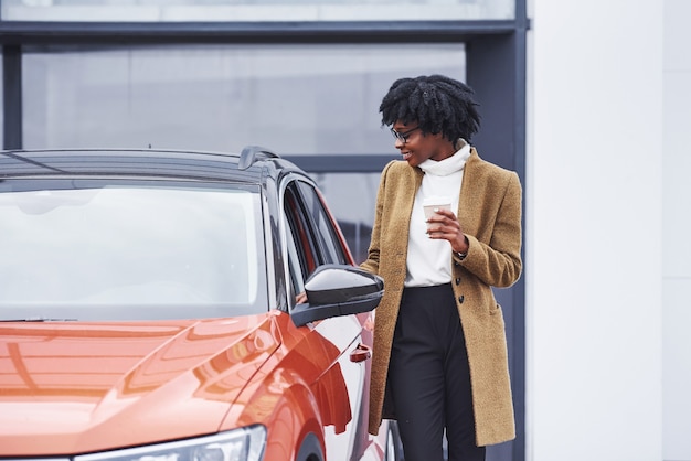 La giovane donna afroamericana in vetri e con la tazza della bevanda sta all'aperto vicino all'automobile moderna.