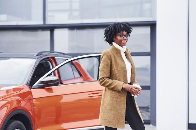 La giovane donna afroamericana in vetri e con la tazza della bevanda sta all'aperto vicino all'automobile moderna.