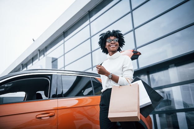 La giovane donna afroamericana in vetri sta all'aperto vicino all'automobile moderna dopo lo shopping con i pacchetti.