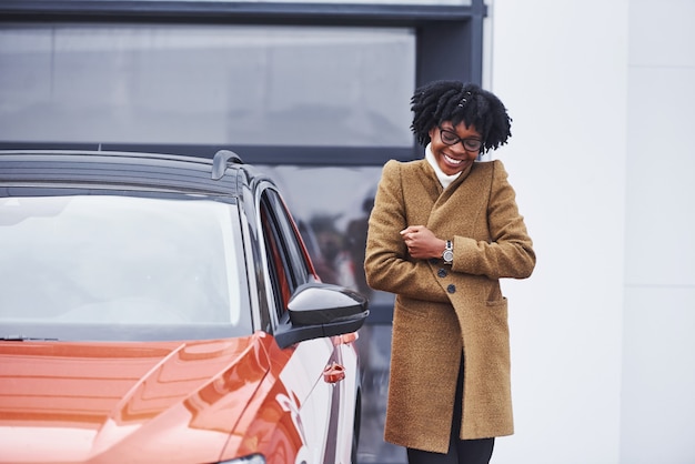 La giovane donna afroamericana in vetri sta all'aperto vicino all'automobile moderna.