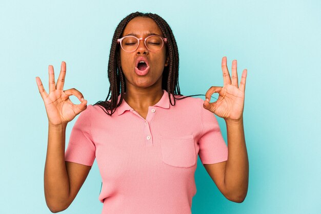 La giovane donna afroamericana isolata su fondo blu si rilassa dopo una dura giornata di lavoro, sta eseguendo lo yoga.