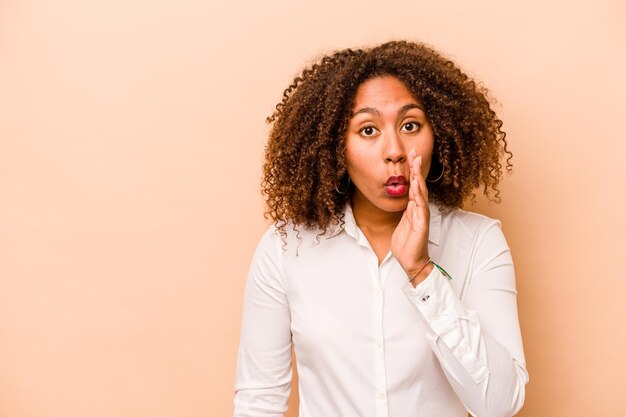 La giovane donna afroamericana isolata su sfondo beige sta dicendo una notizia segreta di frenata calda e sta guardando da parte