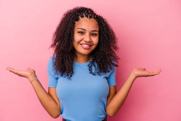 La giovane donna afroamericana isolata su sfondo rosa fa la scala con le braccia, si sente felice e sicura di sé.