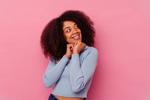 La giovane donna afroamericana isolata sul rosa tiene le mani sotto il mento, sta guardando felicemente da parte.