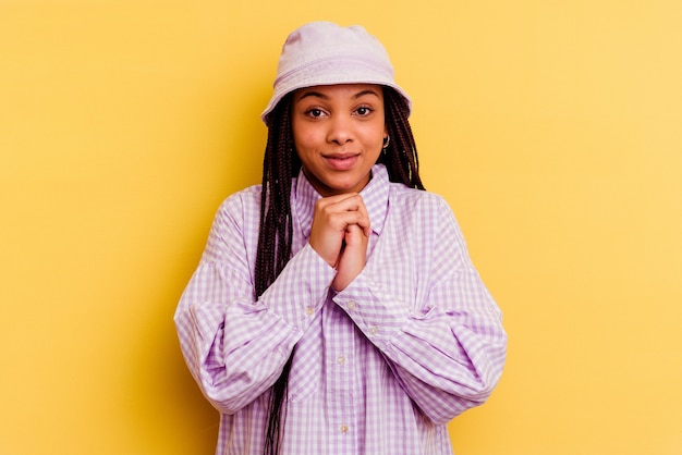 La giovane donna afroamericana isolata sulla parete gialla tiene le mani sotto il mento, sta guardando felicemente da parte.