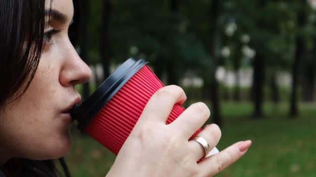 La giovane donna alla moda beve il caffè caldo di mattina in tazza di carta rossa di eco all'aperto nel parco di autunno. Vista laterale del primo piano della tazza di caffè da asporto della tenuta della giovane donna.