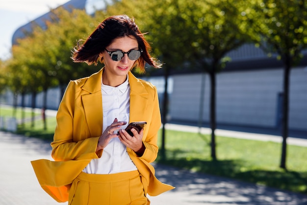 La giovane donna alla moda di affari utilizza lo smartphone mentre cammina vicino al centro di affari