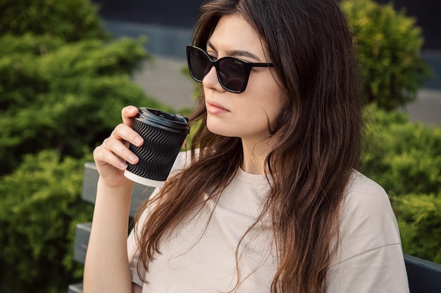 La giovane donna alla moda in occhiali da sole si siede su una panchina e beve il caffè