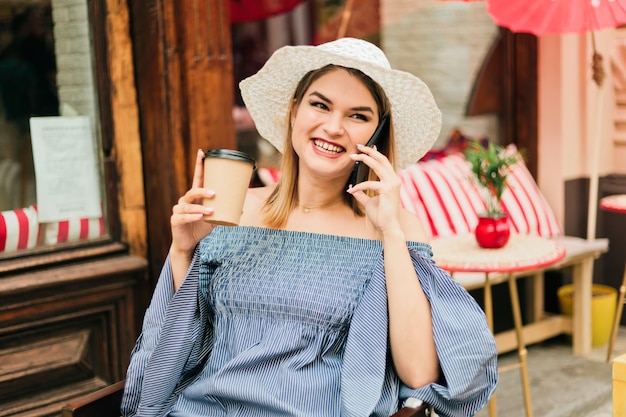 La giovane donna alla moda in un cappello e vestito si siede al caffè all'aperto e beve il caffè mentre parla al telefono