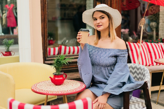 La giovane donna alla moda in un cappello e vestito si siede al caffè all'aperto e beve il caffè