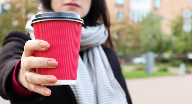 La giovane donna alla moda in un cappotto e una sciarpa beve il caffè caldo di mattina in un bicchiere di carta eco rosso all'aperto in un parco autunnale. Primo piano di una giovane donna che tiene una tazza di caffè da asporto, profondità di campo ridotta.