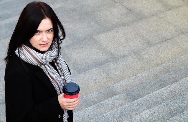 La giovane donna alla moda in un cappotto e una sciarpa beve il caffè caldo di mattina in un bicchiere di carta eco rosso all'aperto in un parco autunnale. Ritratto di una giovane donna che tiene una tazza di caffè da asporto, profondità di campo ridotta.