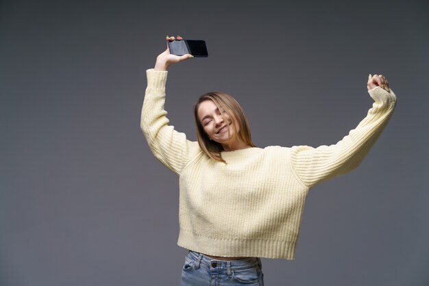 La giovane donna allegra con un maglione giallo su un muro grigio sta ballando con un telefono in mano