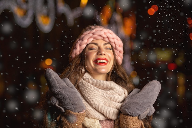La giovane donna allegra indossa cappello rosa, guanti e sciarpa che cammina in città la sera durante la nevicata