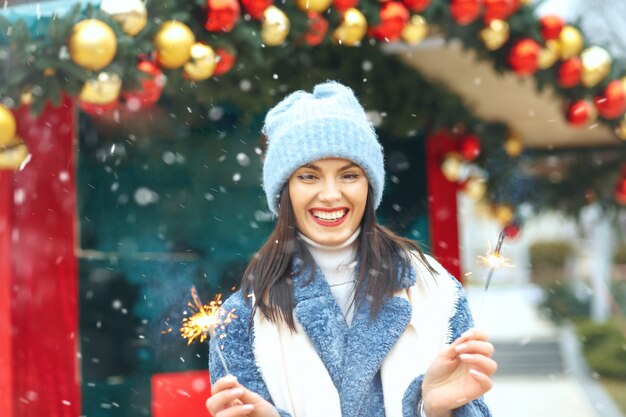 La giovane donna allegra indossa un cappotto blu che si gode le vacanze con le luci del Bengala durante la nevicata