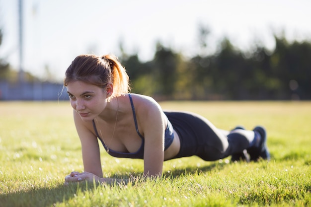 La giovane donna allegra negli sport copre l'addestramento nel campo all'alba. Ragazza che sta nella posizione della plancia sull'erba in un parco della città.