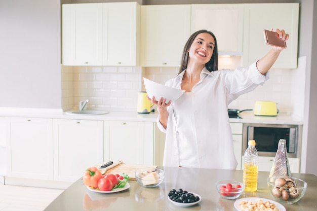 La giovane donna allegra sta in cucina e tiene la ciotola con insalata. Registra video e sorrisi. La ragazza sembra felice.