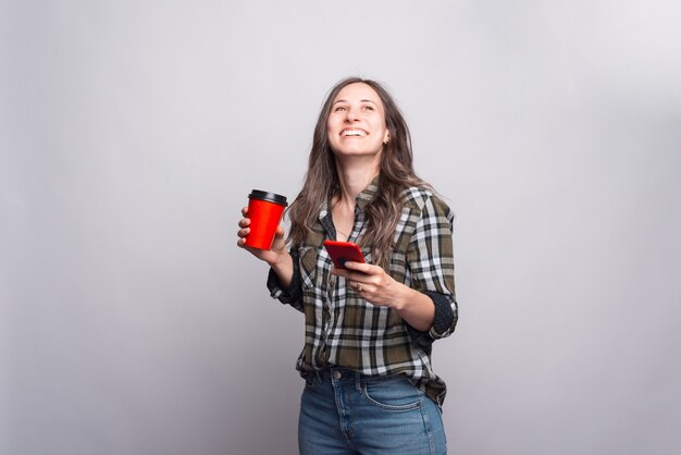 La giovane donna allegra sta tenendo una tazza con una bevanda calda e un telefono vicino al muro grigio