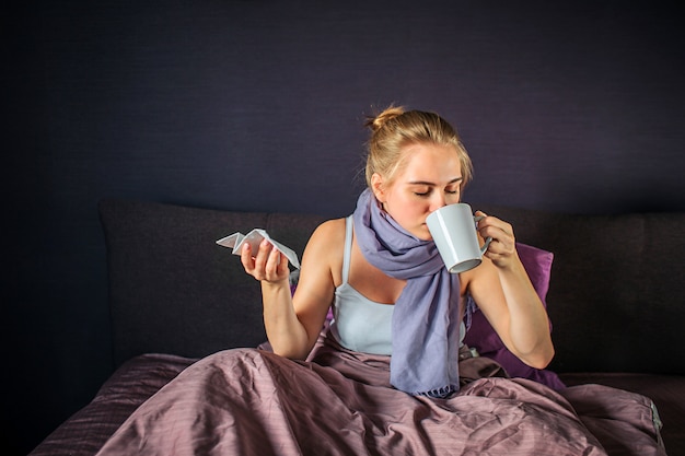 La giovane donna ammalata si siede sul letto e beve dalla tazza bianca. Tiene il tessuto secco in un'altra mano. Le sue gambe sono coperte di coperta.