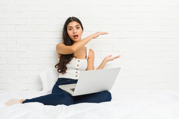 La giovane donna araba che lavora con il suo computer portatile sul letto ha colpito e stupito tenendo uno spazio della copia fra le mani.