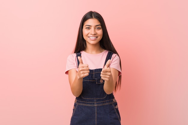 La giovane donna araba graziosa che indossa un denim dei jeans con i pollici aumenta, incoraggia qualcosa, sostiene e rispetta il concetto.