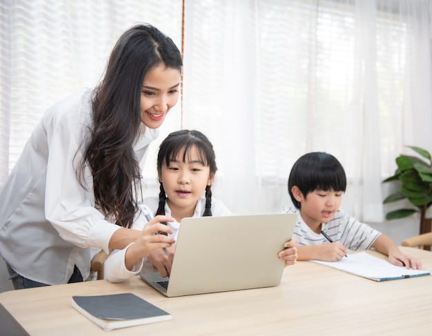 La giovane donna asiatica aiuta suo figlio a fare i compiti con la figlia che per mezzo del computer portatile si siede accanto sulla tavola in salone a casa.