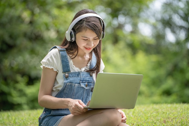 La giovane donna asiatica attraente indossa l'auricolare per ascoltare lo studio di sviluppo del corso online digitale online nel parco