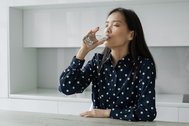 La giovane donna asiatica beve l'acqua da un bicchiere seduta in cucina una giovane donna sorride mentre