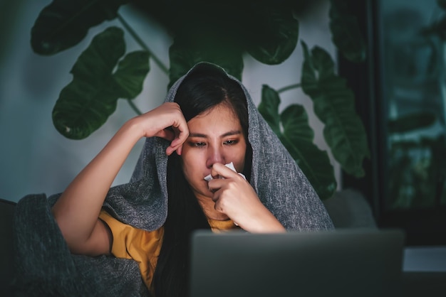 La giovane donna asiatica che soffia il naso e starnutisce nella carta velina mentre lavora con il computer portatile di notte sperimenta il concetto di assistenza sanitaria di sintomi di allergia e influenza