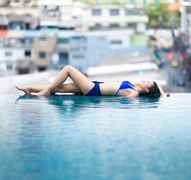 La giovane donna asiatica degli anni '20 dorme lungo il bordo dell'orizzonte della piscina con vista sulla città del cielo blu