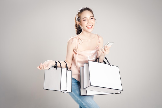 La giovane donna asiatica è felice con le borse della spesa e usa il cellulare in mano su sfondo grigio