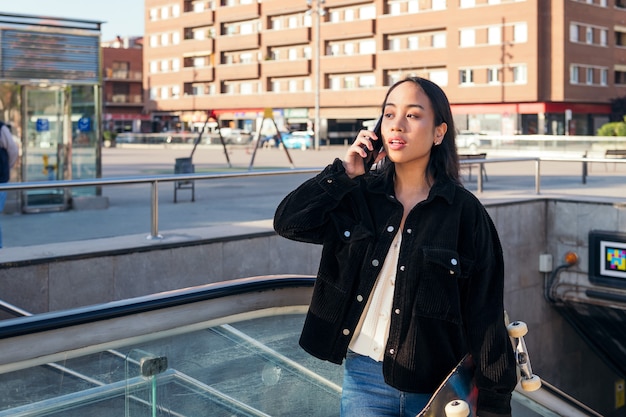 La giovane donna asiatica esce dalla stazione della metropolitana con lo skateboard in mano mentre parla al telefono