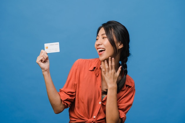 La giovane donna asiatica mostra la carta di credito con espressione positiva, sorride ampiamente, vestita con abiti casual sentendo felicità e stando isolata sulla parete blu. Concetto di espressione facciale.