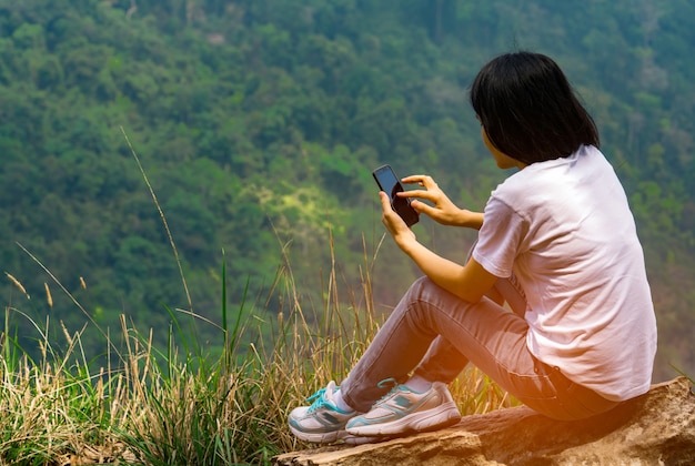 La giovane donna asiatica si siede sulla roccia alla scogliera e sta usando lo Smart Phone