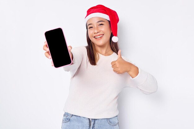 La giovane donna asiatica sorridente in un cappello di Natale che mostra il telefono cellulare con schermo vuoto fa il pollice sul gesto raccomandando l'app mobile isolata su sfondo bianco
