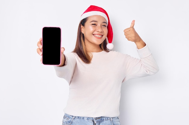 La giovane donna asiatica sorridente in un cappello di Natale che mostra il telefono cellulare con schermo vuoto fa il pollice sul gesto raccomandando l'app mobile isolata su sfondo bianco