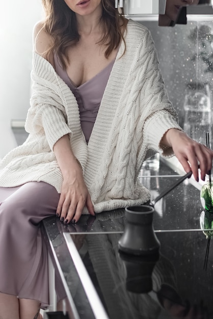 La giovane donna attraente che porta il maglione bianco tricottato si siede sul ripiano del tavolo della cucina che produce il caffè preparato di mattina