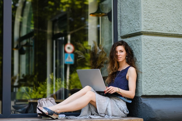 La giovane donna attraente con il computer portatile si siede sul davanzale all'aperto