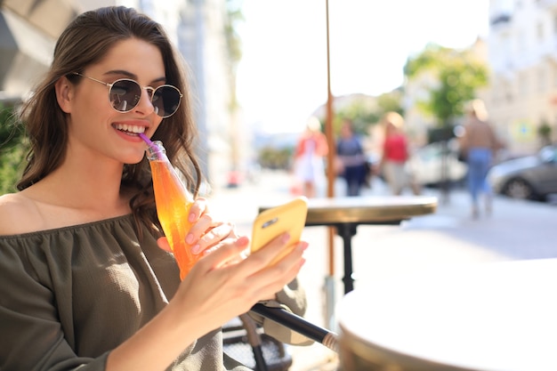 La giovane donna attraente in occhiali da sole tiene una bottiglia con succo, facendo uso dello smartphone, all'aperto.