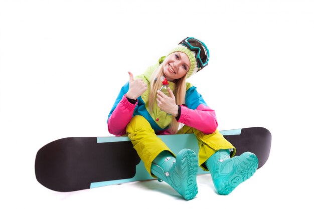 La giovane donna attraente in vestito di sci e vetri di sci si siede sullo snowboard, tiene l'acqua