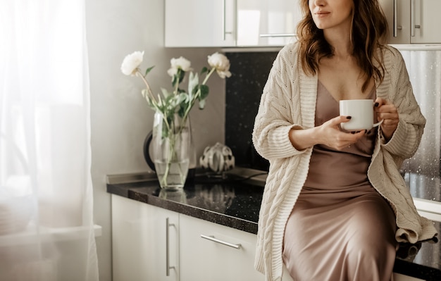 La giovane donna attraente si siede sul ripiano del tavolo che tiene la tazza di caffè caldo nella stanza della cucina con l'interno moderno bianco