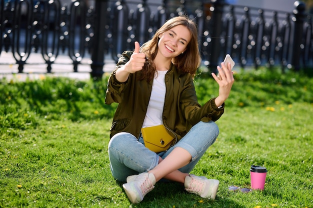 La giovane donna attraente sorridente si rilassa nel parco pubblico che si siede sull'erba utilizza lo smartphone e mostra il pollice sul gesto